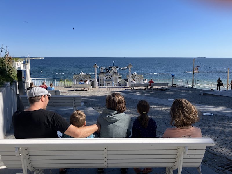 5 Personen, davon 2 Erwachsene und 3 Kinder sitzen auf einer Bank und schauen auf das blaue Meer.