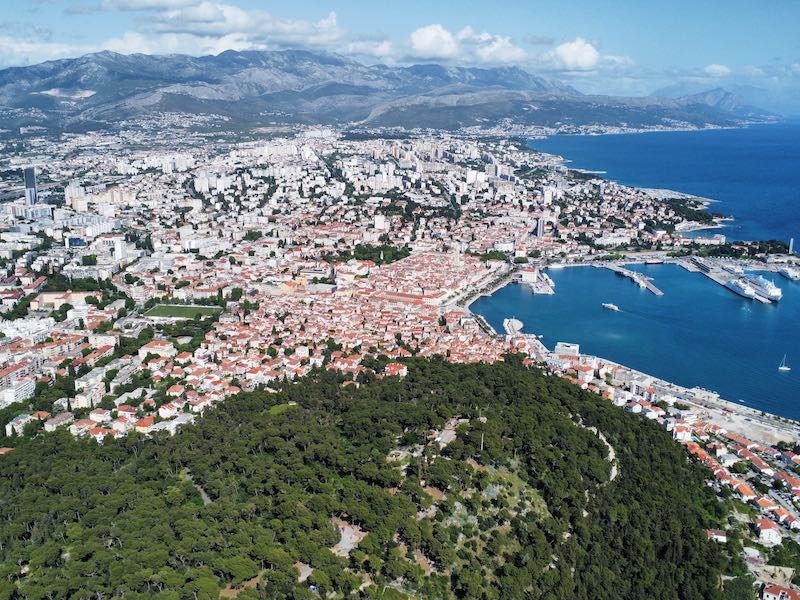 Blick vom Flugzeug auf die Stadt Split in Dalmatien am Meer, im Hintergrund ist ein Gebirgsmassiv zu sehen.
