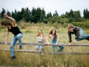 Vier Teenager (2 Mädchen und 2 Jungs) laufen über eine Wiese. Die beiden Jungs springen über einen Zaun.