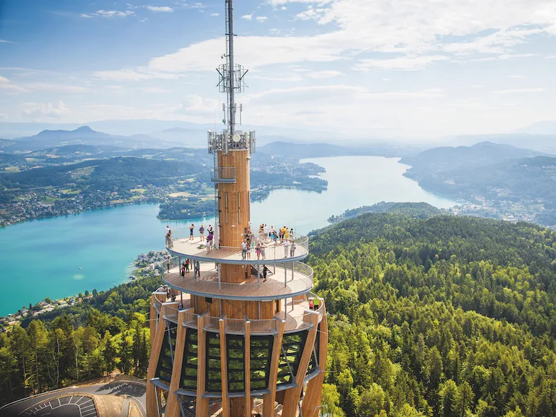 Ein Aussichtsturm mit Besuchern, im Hintergrund ist ein See und es sind Berge zu sehen.